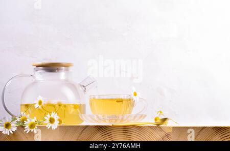 Transparente Glas-Teekanne und -Tasse mit gebrühtem gesundem Kamillentee, Kamillenblüten auf Holztisch auf hellem neutralem Hintergrund. Kopierbereich. Kräuter Stockfoto