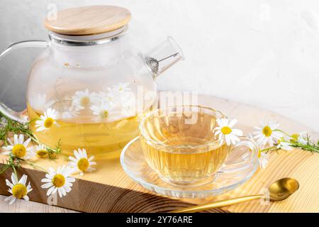 Teetrinken für die Gesundheit. Teekanne aus Glas und Tasse mit gebrühtem gesundem Kamillentee, goldenem Löffel, Kamillenblüten auf Holzbrett. Nahaufnahme. Kräutermedizin Stockfoto