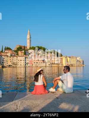 Ein Paar sitzt auf einem Steinpier und bestaunt die malerischen Hügel von Rovinj. Das warme Leuchten des Sonnenuntergangs färbt den Himmel, reflektiert vom ruhigen Wasser und schafft einen Moment der Ruhe. Stockfoto