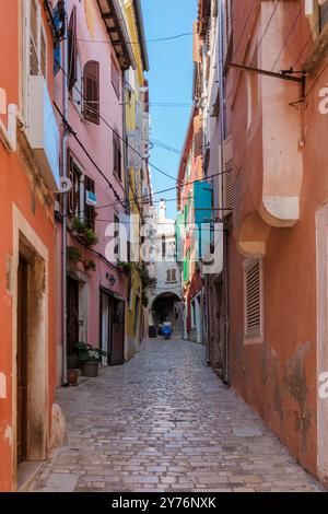 Im Herzen von Rovinj, Kroatien, bietet eine enge Kopfsteinpflasterstraße einen Blick auf lebhafte Fassaden und einladende Eingänge. Die malerische Gasse ist von Sonnenlicht umgeben und zeigt den Charme dieser Küstenstadt. Stockfoto