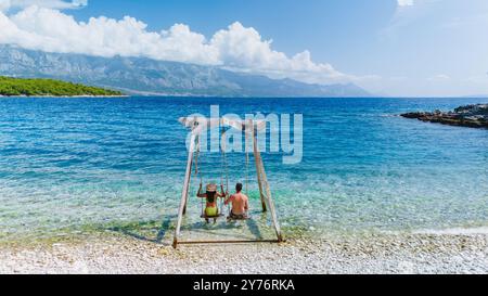 An einem sonnigen Tag auf Brac Island genießen zwei Personen eine Schaukel am kristallklaren Wasser, umgeben von atemberaubenden Bergen. Lachen erfüllt die Luft, während sie diesen wunderschönen Urlaub am Meer schätzen. Stockfoto