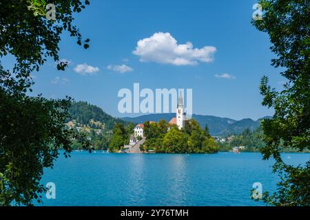 Ein ruhiger Sommertag am Lake Bled bietet eine malerische Insel mit einer malerischen Kirche, umgeben von lebhaftem Grün und ruhigem türkisfarbenem Wasser, die Besucher einlädt, ihre Schönheit zu erkunden. Stockfoto