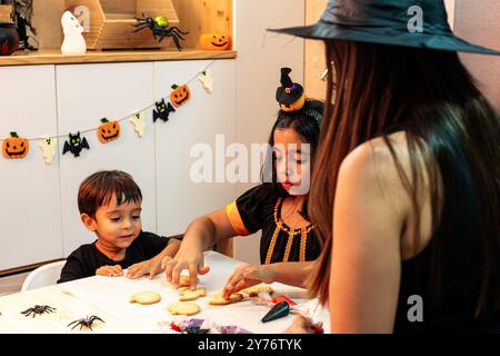 Eine Frau hilft zwei Kindern, Halloween-Kekse zu machen. Die Szene ist festlich und lustig, da die Kinder in Halloween-Kostümen verkleidet sind und die Frau i Stockfoto