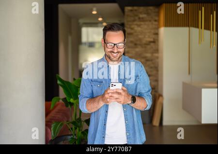 Positiver männlicher Manager in Brille, der Social-Media-Anwendungen über Handy im modernen Büro überprüft Stockfoto