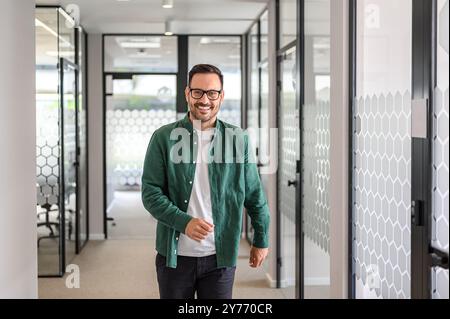 Porträt eines inspirierten männlichen Unternehmers mit Brille, der selbstbewusst im Bürokorridor lächelt und geht Stockfoto