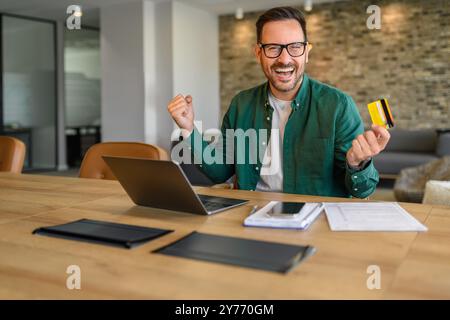 Fröhlicher Geschäftsmann, der vor Freude schreiend und Fäuste schüttelt, während er online über einen Laptop am Schreibtisch im Büro bezahlt Stockfoto