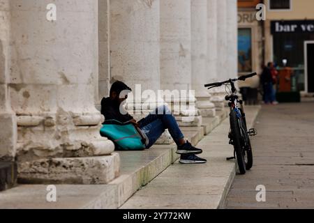 Vicenza, Italien - 13. September 2024: Ein Arbeiter aus Deliveroo macht eine Pause, sitzt auf der Treppe und lehnt sich an seine Arbeitstasche, während sein Fahrrad ruht Stockfoto