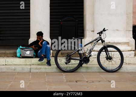 Vicenza, Italien - 13. September 2024: Ein Arbeiter aus Deliveroo macht eine Pause, sitzt auf der Treppe und lehnt sich an seine Arbeitstasche, während sein Fahrrad ruht Stockfoto