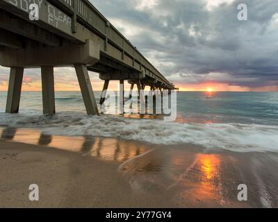 Bewölkter Sonnenaufgang am Deerfield Beach Pier, Florida, wenn das Sonnenlicht durch die Wolken über den Wellen und der Küste bricht Stockfoto