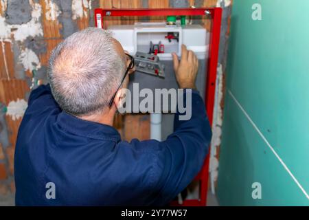 Ein ausgereifter Klempner, der den neuen WC-Unterputzrahmen für die wandmontierte Toilette installiert. Bauherren Wohnungsbau- oder Renovierungsarbeiten. Stockfoto