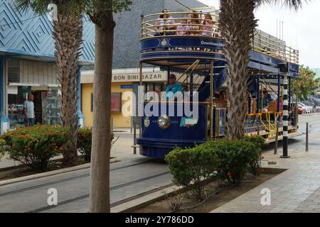 Der Trolley in der Innenstadt ist voll von Touristen, die durch eine enge Straße mit farbenfrohen Gebäuden und Palmen auf der Karibikinsel Aruba in Oranjestad fahren. Stockfoto