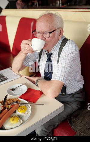 Ein älterer Mann hält eine Tasse Kaffee mit einem Frühstücksteller auf dem Tisch, trägt eine Brille und ein gestreiftes Hemd mit Krawatte, sitzt in einem roten Diner, liest Zeitung und denkt nach Stockfoto