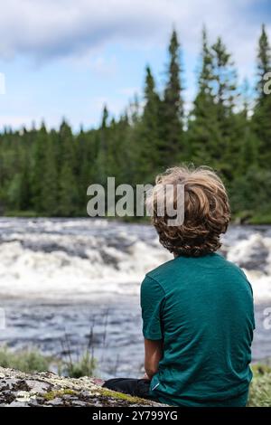 Rückansicht eines kleinen Jungen, der an einem Sommertag in der Nordic Wilderness auf einem felsigen Flussufer sitzt und über die rauschenden Stromschnellen blickt Stockfoto