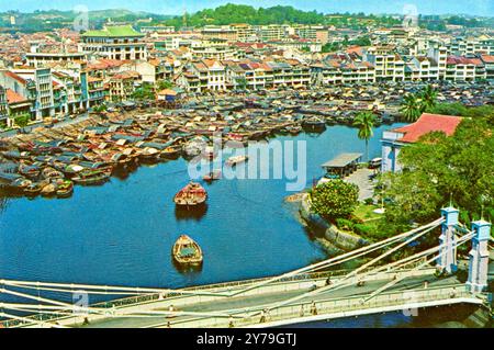 Ein Postkartenbild aus den 1960er Jahren des Boat Quay und der Cavenagh Bridge, das Sampanboote zeigt, die Lebensmittel und Fracht auf und ab den Singapore River transportierten. Aus diesem Blickwinkel ist es möglich, dass das Bild aus dem heutigen Fullerton Hotel, dem ehemaligen General Post Office und anderen Regierungsbehörden stammt. Heute sind die Boote alle weg, neben ein paar Touristenbooten, ebenso das Gebäude und der Parkplatz unten rechts. Die Uferlinie besteht heute aus Hochhäusern, aber viele der kleinen Ladenhäuser (Shoptop) sind erhalten und werden als Restaurants und Bars genutzt Stockfoto