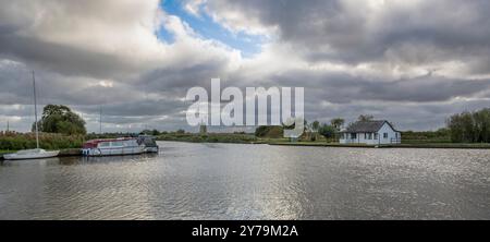 Ein Blick auf die norfolk Broads in Thurne. Das Foto zeigt ein kleines Haus am Fluss und eine Windmühle in der Ferne. Kleine Boote liegen vor Stockfoto