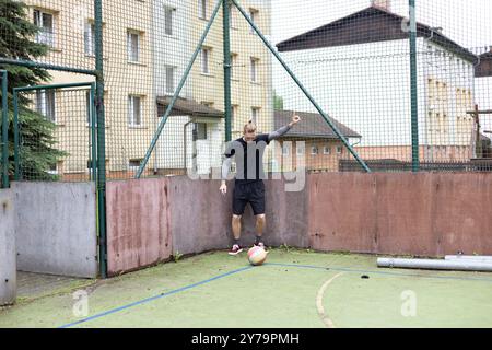Ein junger Mann steht auf einem Freigelände gegen einen Zaun und signalisiert mit der Hand, während ein Fußball nahe seinem Fuß liegt. Die Szene kombiniert einen Freizeitsport Stockfoto