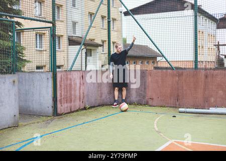Ein junger Mann steht auf einem Freigelände gegen einen Zaun und signalisiert mit der Hand, während ein Fußball nahe seinem Fuß liegt. Die Szene kombiniert einen Freizeitsport Stockfoto