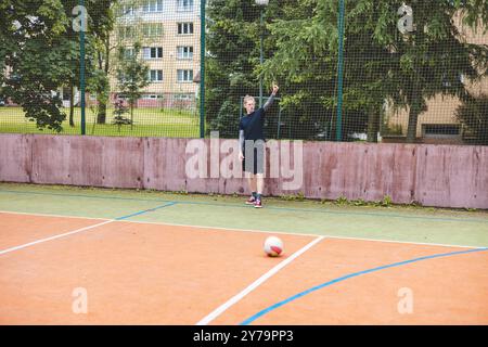 Ein junger Mann steht auf einem Freigelände gegen einen Zaun und signalisiert mit der Hand, während ein Fußball nahe seinem Fuß liegt. Die Szene kombiniert einen Freizeitsport Stockfoto