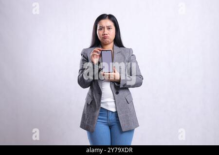 Charme schöne junge Bürofrau indonesien, die zur Kamera beide Hände hält, zeigt Handy-Bildschirm, der grauen Anzug isoliert auf weißem Hintergrund trägt Stockfoto