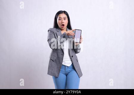 Wunderschönes junges Büromädchen indonesien Charme schockiert zu Kamera Hand seitlich hält zeigt Handy-Bildschirm mit grauem Anzug isoliert auf weißem Rücken Stockfoto