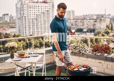 Hübscher junger Mann in lässiger Kleidung, der Barbecue vorbereitet, während er auf der Dachterrasse steht Stockfoto