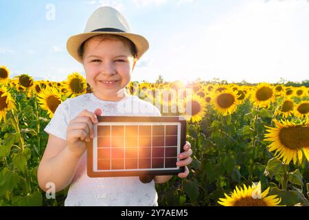 Fröhlich lächelndes Mädchen, das auf einem Feld mit Sonnenblumen steht und ein tragbares Solarmodul in der Hand hält. Alternatives, umweltfreundliches und solares Energiekonzept Stockfoto
