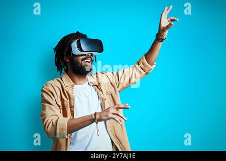 Fröhlicher junger afrikanischer Mann, der eine Virtual-Reality-Brille trägt, während er vor blauem Hintergrund steht Stockfoto