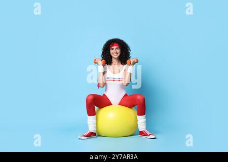 Frau, die aerobe Übungen mit Kurzhanteln und Fitnessball auf hellblauem Hintergrund macht Stockfoto