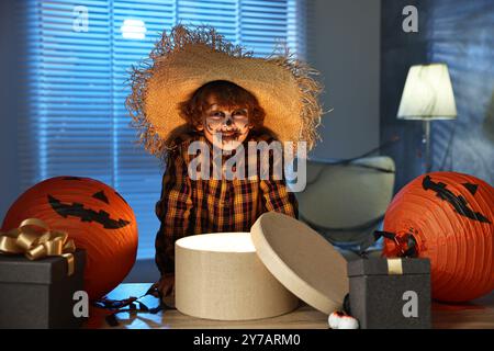 Süßer Junge, der wie Vogelscheuche gekleidet ist, mit festlichem Dekor und Geschenkboxen drinnen in der Nacht. Halloween-Feier Stockfoto