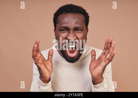 Wütender junger afrikanischer Mann in lässiger Kleidung, der schreiend und in die Kamera blickt, während er vor braunem Hintergrund steht Stockfoto