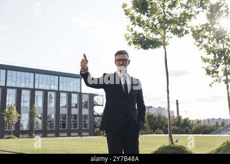 Selbstbewusster reifer Geschäftsmann mit Brille, der im Freien seinen Daumen nach oben zeigt Stockfoto