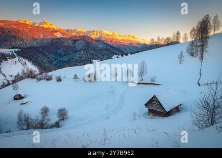 Bewundernswerte Winterlandschaft mit schneebedeckter Holzhütte am Hang und wunderschönen hohen Bergen im Hintergrund bei Sonnenuntergang, Bucegi-Berge, Karpaten, Rom Stockfoto