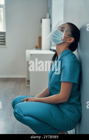 Traurige junge Krankenschwester in schützender Arbeitskleidung, die während der Arbeit im Krankenhaus die Augen geschlossen hält Stockfoto