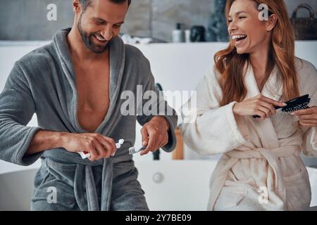 Liebevolles junges Paar in Bademänteln, lacht während der morgendlichen Routine Stockfoto
