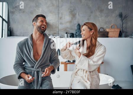 Liebevolles junges Paar in Bademänteln, lacht während der morgendlichen Routine Stockfoto