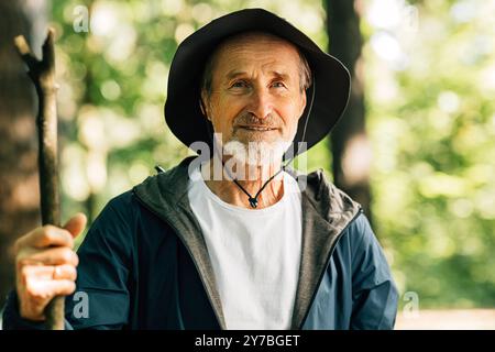 Porträt eines älteren Mannes, der einen Hut trägt und einen Stock hält. Reifer Mann, der während seines Waldgangs in die Kamera schaut. Porträt eines älteren männlichen Trägers Stockfoto