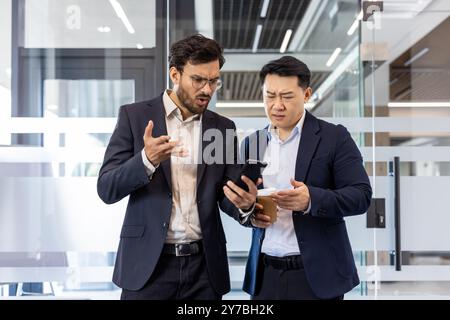 Zwei Geschäftsleute in modernen Büroumgebungen diskutieren intensiv über Nachrichten, die auf dem Smartphone angezeigt werden. Kollegen in beruflicher Kleidung äußern Bedenken und betonen Teamarbeit und Dringlichkeit in der Geschäftskommunikation Stockfoto