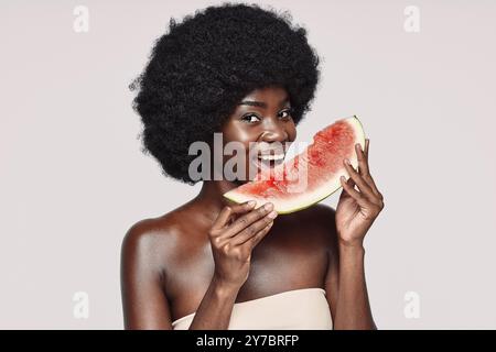Porträt einer schönen jungen afrikanischen Frau, die ein Stück Wassermelone hält und lächelnd vor grauem Hintergrund steht Stockfoto