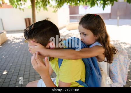 Junge Mädchen mit Rucksack bedeckt spielerisch die Augen des Jungen, während sie draußen sitzen. Fängt Kindheit, Freundschaft und Spaß ein. Stockfoto