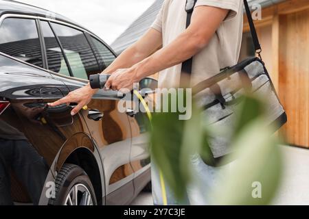 Mann, der eine Steckdose öffnet und ein Ladegerät in ein schwarzes Elektroauto an eine EV-Ladestation in der Stadtstraße stellt Stockfoto