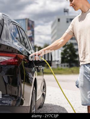 Mann, der eine Steckdose öffnet und ein Ladegerät in ein schwarzes Elektroauto an eine EV-Ladestation in der Stadtstraße stellt Stockfoto