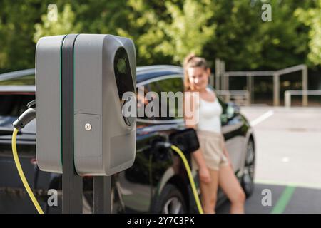 Lächelndes Mädchen, das das Ladegerät in ihr schwarzes Elektroauto steckte und es mit der auf dem Parkplatz installierten EV-Ladestation verband Stockfoto