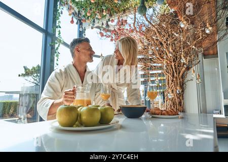 Wunderschönes reifes Paar in Bademänteln, das zusammen frühstückt, während es Zeit in der Küche verbringt Stockfoto