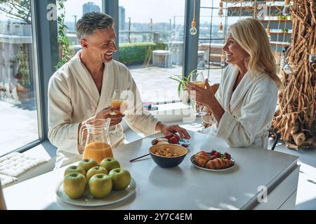 Wunderschönes reifes Paar in Bademänteln, das zusammen frühstückt, während es Zeit in der Küche verbringt Stockfoto