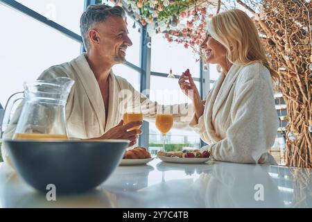 Wunderschönes reifes Paar in Bademänteln, das zusammen frühstückt, während es Zeit in der Küche verbringt Stockfoto