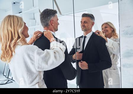 Schöne reife Frau, die den Anzug ihres Mannes anpasst, während beide zu Hause vor dem Spiegel stehen Stockfoto