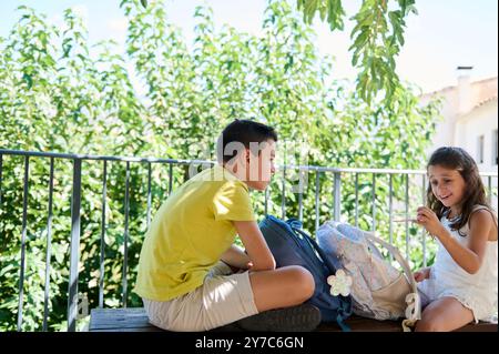 Zwei Kinder sitzen auf einer Bank mit Rucksäcken und genießen einen sonnigen Tag draußen. Perfekte Darstellung von Kindheitsfreude und Freundschaft in einer natürlichen Umgebung. Stockfoto