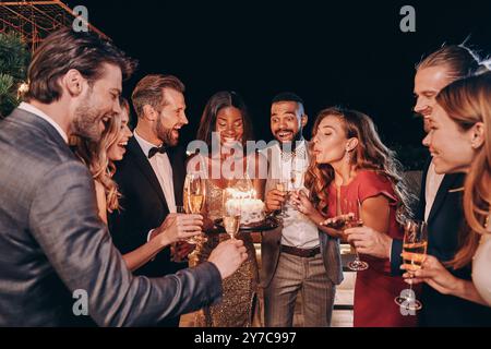 Schöne junge Frau im Abendkleid, die Kerzen auf Kuchen bläst, während sie mit Freunden Geburtstag feiert Stockfoto