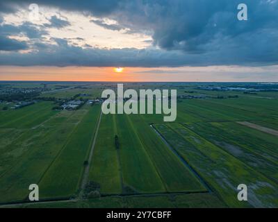Blick aus der Vogelperspektive auf grüne landwirtschaftliche Felder und ein kleines Dorf vor bewölktem Himmel bei Sonnenuntergang. Niederländische Landschaft. Stockfoto