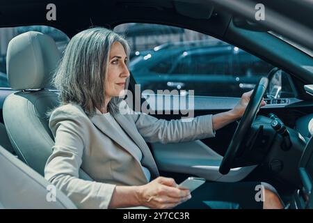 Reife schöne Frau in smarter Freizeitkleidung lächelt und benutzt das Telefon während des Autofahrens Stockfoto